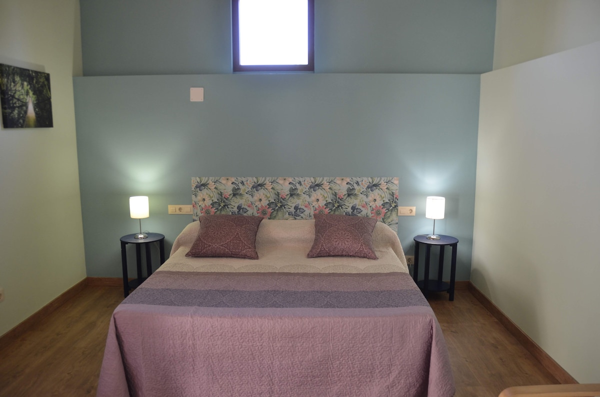 A spacious sleeping area is presented with a floral headboard and soft bedding. Two bedside tables with lamps frame the bed, providing soft illumination. A window above allows natural light to filter in, highlighting the calming color palette of the room.
