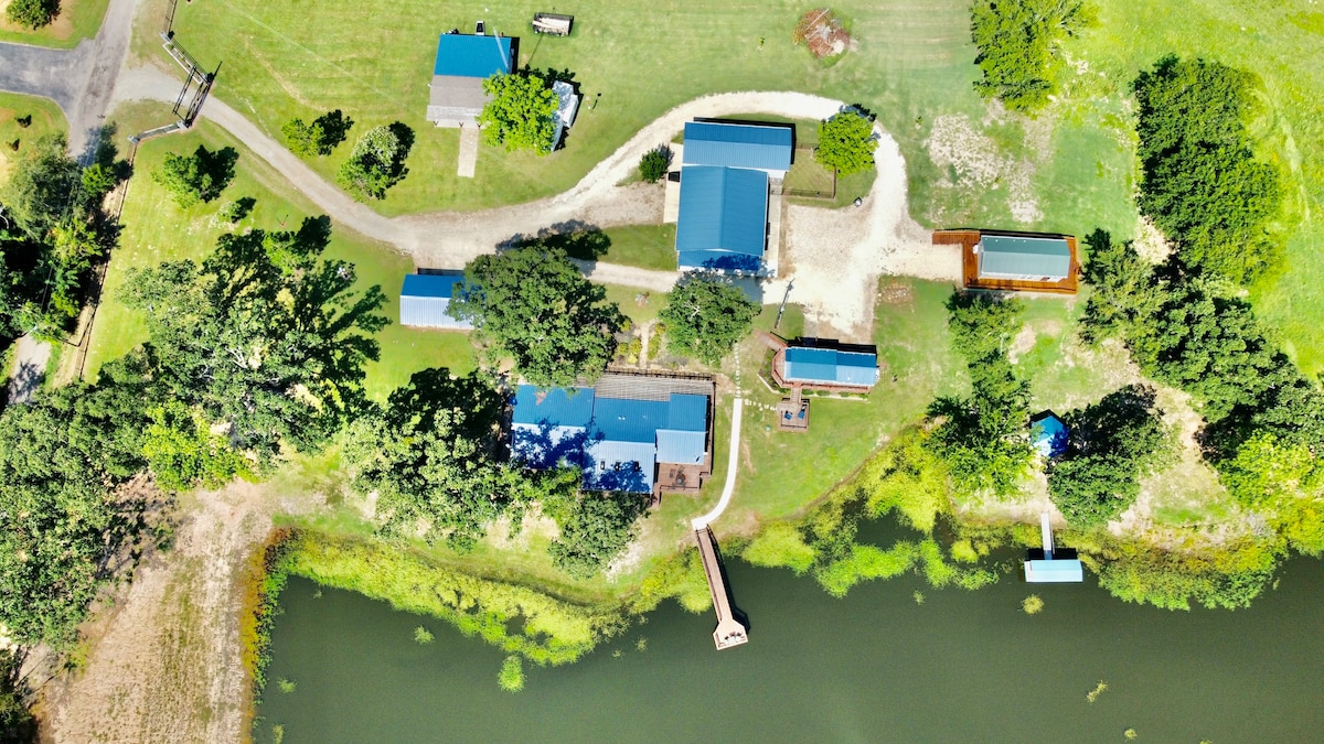 An aerial view captures a serene landscape featuring multiple buildings set on lush green grounds. A winding path leads to a private lake, with a dock extending into the water. Surrounding trees provide shade and enhance the tranquil environment.