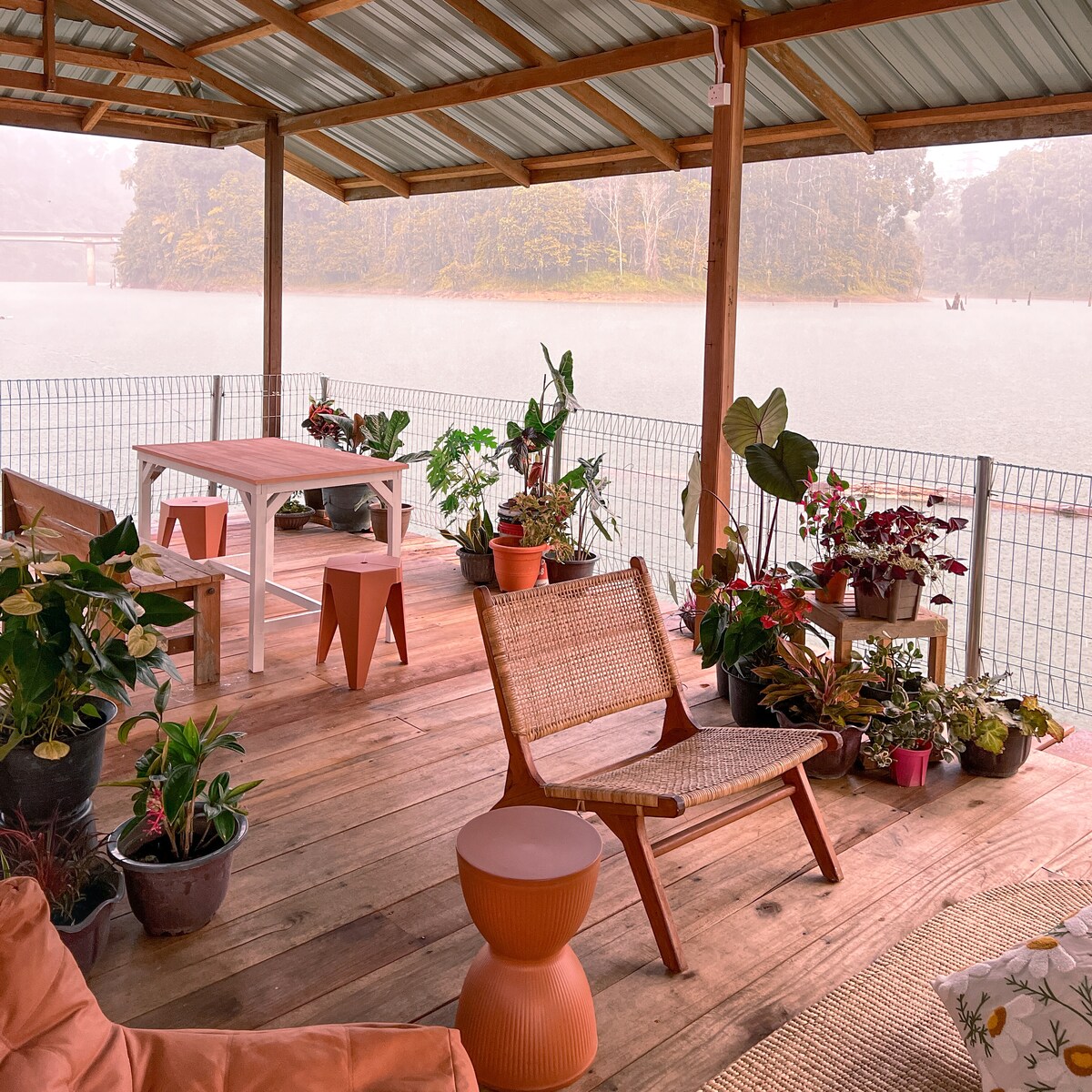 A covered outdoor seating area features a wooden table and several chairs, surrounded by potted plants. The floor is made of wooden planks, and a tranquil water view is visible in the background, creating a serene atmosphere.