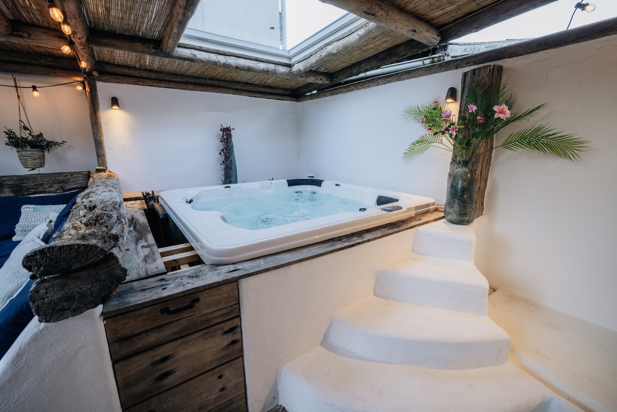 A serene indoor space features a sunken jacuzzi illuminated by natural light from an overhead skylight. Wooden accents enhance the rustic vibe, while tropical plants add a touch of greenery. Soft white walls and steps lead to the relaxing area.