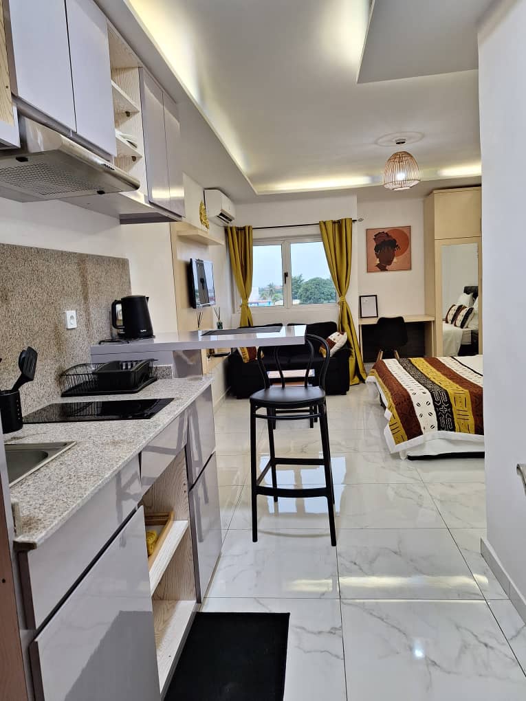 A well-arranged studio apartment features a kitchenette with modern appliances, including a microwave and refrigerator. A workspace with a chair is visible near large windows, while a cozy sleeping area is accented by colorful textiles and wall art, creating a functional living space.