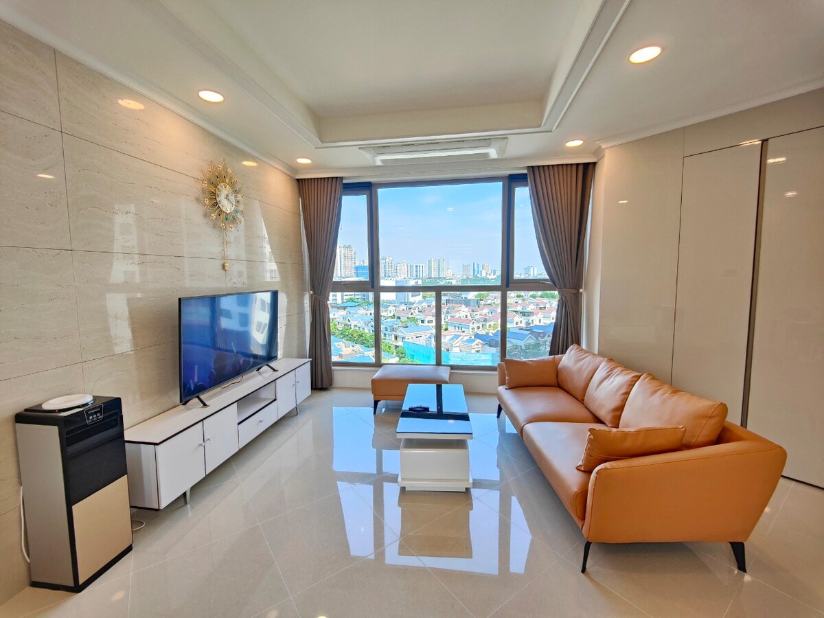 A bright living area features large windows offering a view of the city skyline. A plush, light brown sofa is arranged around a low, white coffee table. A flat-screen TV is mounted on the wall, complemented by neutral decor and polished tile flooring.