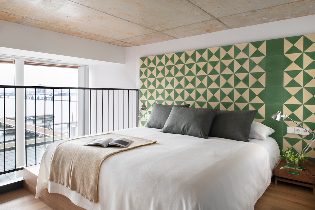 A mezzanine bedroom features a spacious bed with a geometric patterned headboard and neutral bedding. Two large windows allow natural light to fill the room, offering views of the river. A bedside table supports a small plant and an open book.