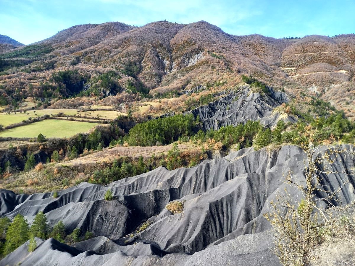 A sweeping landscape features rugged mountain terrain, characterized by sloping gray hills shaped by erosion. Lush green forests line the base of the hills, transitioning into patches of autumn-colored foliage. Open fields are visible in the valley, creating a striking contrast against the mountainous backdrop.