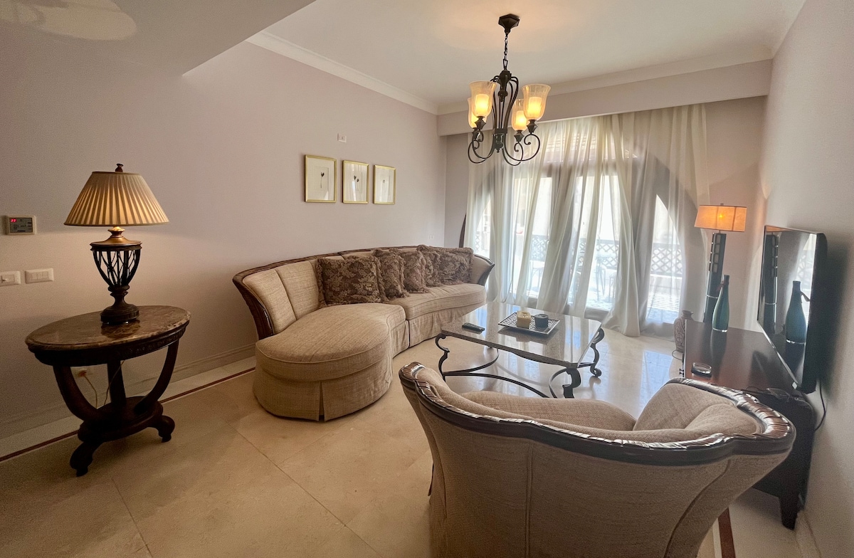 A cozy living room features a curved sofa adorned with decorative cushions, paired with an elegant armchair. A round side table and glass coffee table provide functional surfaces. Soft natural light filters through sheer drapes, creating a warm ambiance in the space.