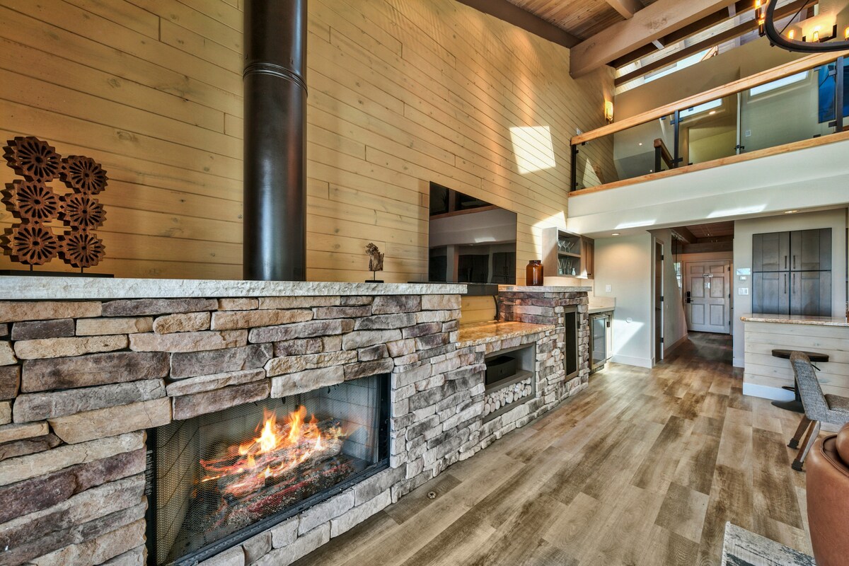 A modern living area features a stone fireplace with an elegant mantel, showcasing a glowing fire. Tall windows allow natural light to illuminate the room, highlighting the wood paneling and open layout that connects to the dining area in the background.