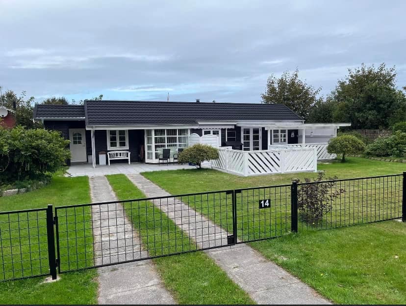 A single-story house is set amidst a green lawn, enclosed by a black fence. A well-maintained pathway leads to the entrance, flanked by shrubs and trees. The front porch is furnished with comfortable seating, inviting relaxation in the outdoor space.