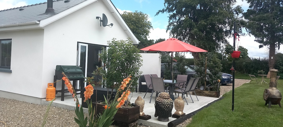 An outdoor patio area features a shaded dining set under a vibrant red umbrella, surrounded by well-maintained gardens. A charcoal grill is positioned nearby, with decorative planters containing blooming flowers adding color to the setting. A gravel pathway leads to the entrance of the cottage.