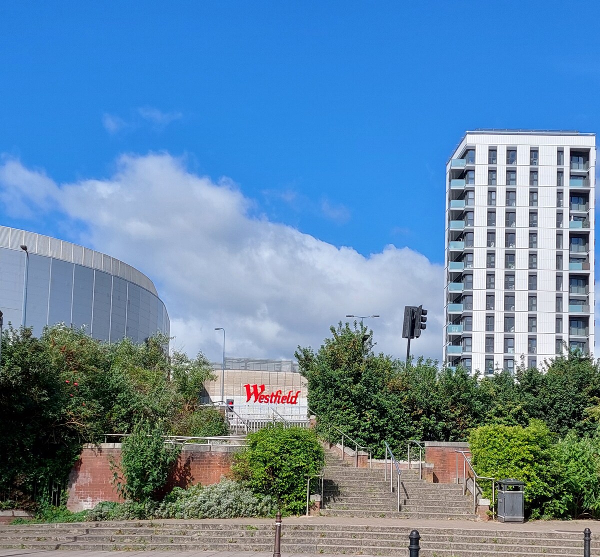 An outdoor view featuring the Westfield shopping centre prominently displayed among greenery. Nearby residential buildings rise in the background under a clear blue sky, suggesting a lively urban atmosphere. Steps lead down towards the entrance, surrounded by well-maintained foliage.