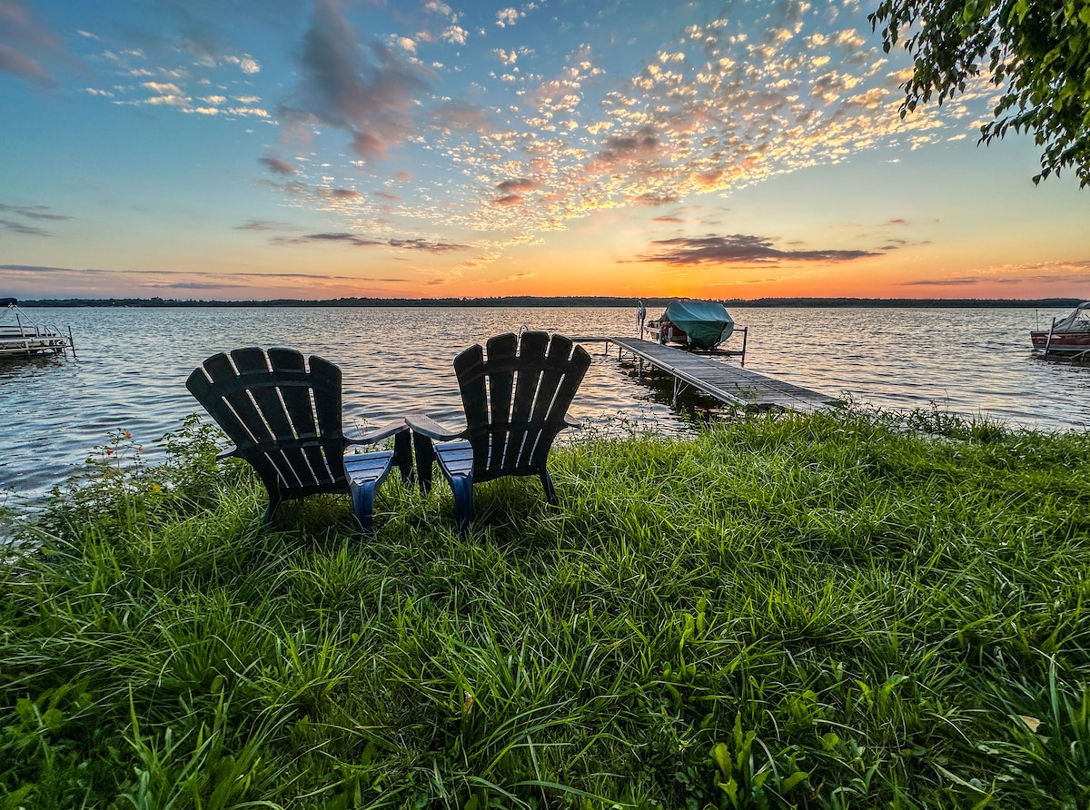 Two Muskoka chairs are positioned on a grassy area facing a tranquil lake at sunset. A dock extends into the water, framed by a colorful sky filled with clouds. The peaceful scene highlights the natural beauty of the waterfront.