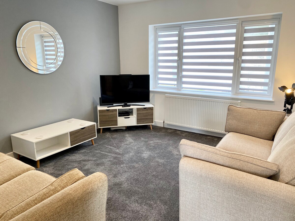 A living room is showcased with a plush carpet and neutral-colored walls. A comfortable sofa is positioned alongside a modern TV stand, which houses a flat-screen television. Large windows with horizontal blinds allow natural light to filter in, complementing the contemporary decor.