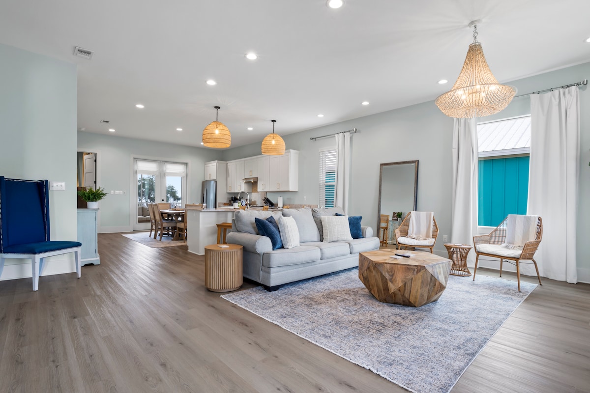 A modern living area features a grey couch facing wicker accent chairs. A wooden coffee table rests on a soft area rug. Natural light floods through large windows, while light fixtures hang from the ceiling, complementing the open layout and coastal-inspired décor.