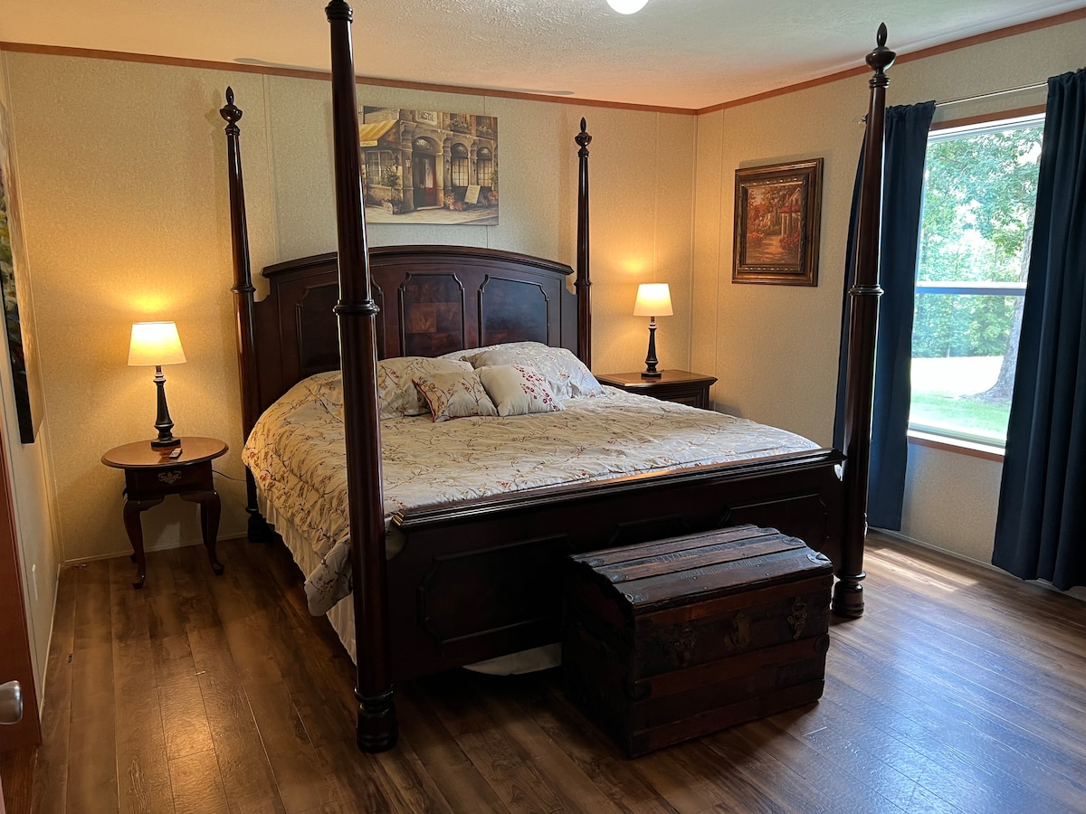 A spacious bedroom features a four-poster bed with a floral patterned comforter. Two bedside lamps illuminate wooden nightstands on either side. A decorative trunk is placed at the foot of the bed, and a large window allows natural light to brighten the room.