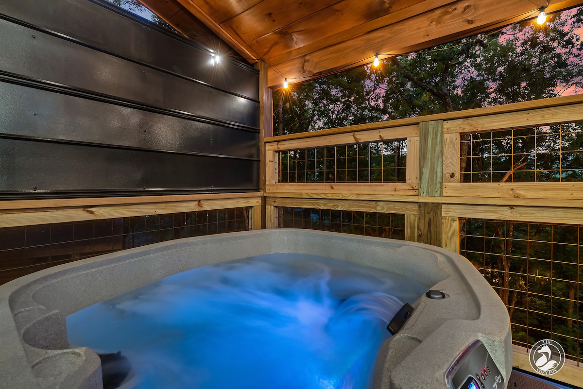 A hot tub is situated on a covered deck, surrounded by wooden railings. Warm blue lights illuminate the water, creating a calming ambiance. A backdrop of trees is visible, with soft natural light filtering through, enhancing the tranquil outdoor setting.