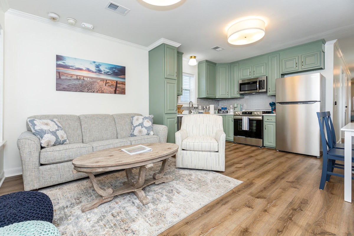 The open living space features a light-colored sofa and a comfortable armchair facing a rustic wooden coffee table. A well-equipped kitchen is visible in the background, complemented by green cabinetry and stainless steel appliances. A beach-themed artwork adorns the wall, enhancing the coastal ambiance.