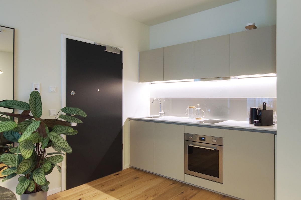 A modern kitchen area is depicted, featuring sleek cabinetry and a subtle color palette. A built-in oven and a sink are visible, complemented by a kettle. Natural wood flooring contrasts with the smooth surfaces, while a potted plant adds a touch of greenery.