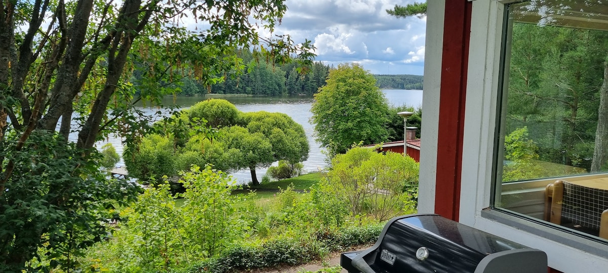 A gas grill sits on a patio overlooking a serene lake, surrounded by lush greenery and trees. The scene captures the tranquil atmosphere, with soft clouds visible in the sky above the water, creating a peaceful outdoor setting.