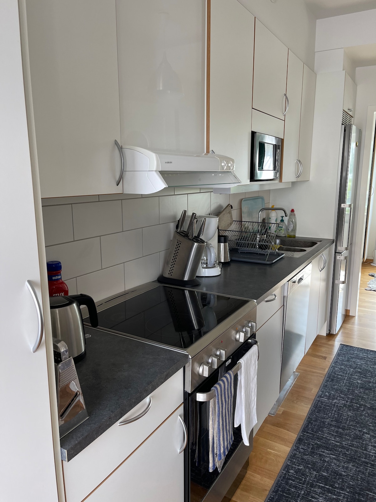 A modern kitchen is equipped with a sleek countertop, featuring a stovetop, electric kettle, and kitchen appliances. A stainless steel refrigerator stands to the side, while dishware and utensils are neatly organized on the counter. Light-toned cabinetry provides ample storage space.