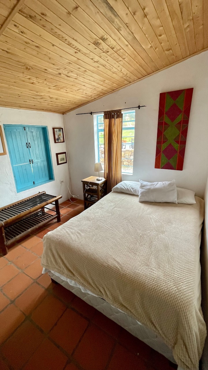 Secondary room, rustic and harmonious. It has a double bed, bedding, wooden bedside tables, and a sample of local artists. 