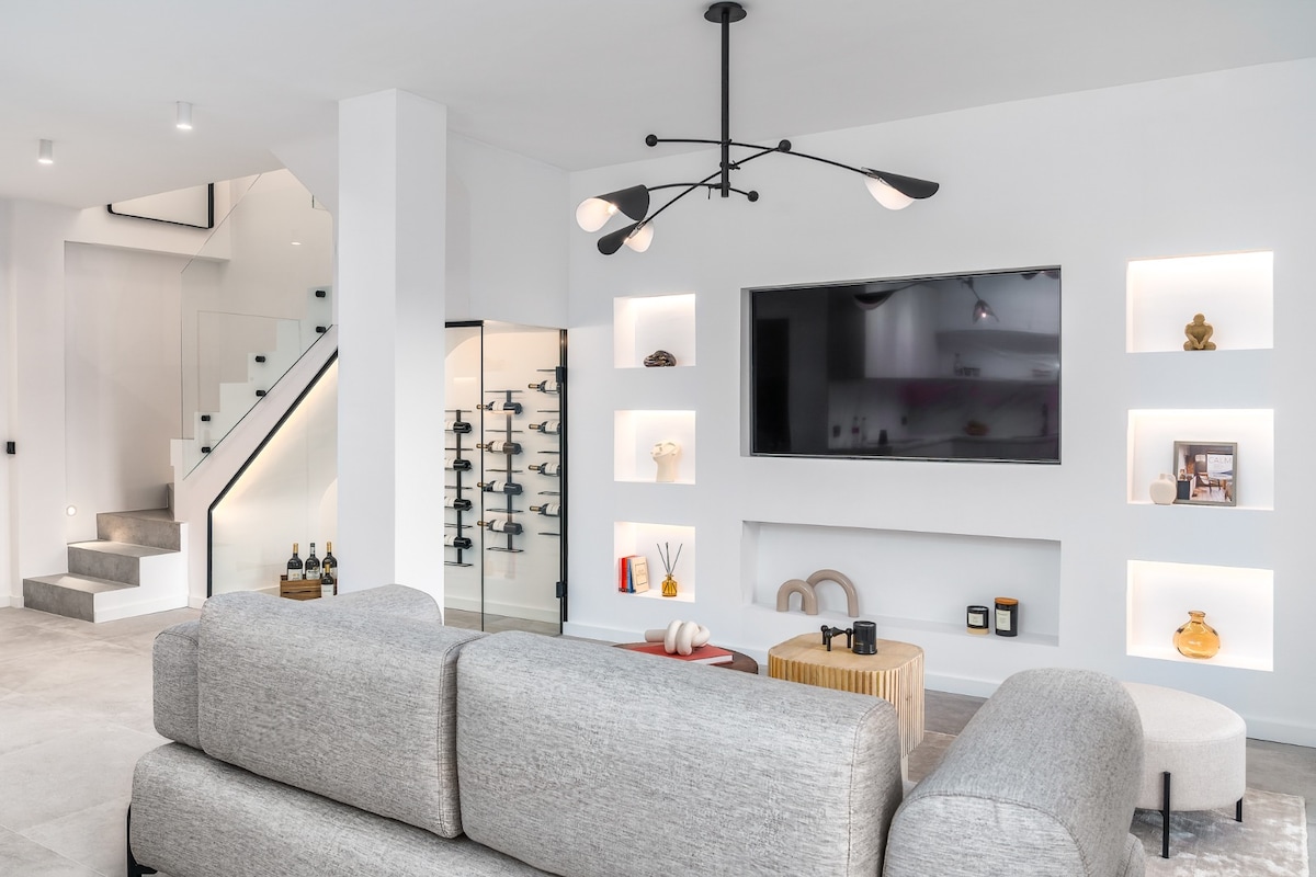A modern living area is showcased, featuring a light gray sectional sofa facing a wall-mounted television. White built-in shelves display decorative items, and ambient lighting is provided by a sleek light fixture. A staircase with a glass railing leads to the upper level.