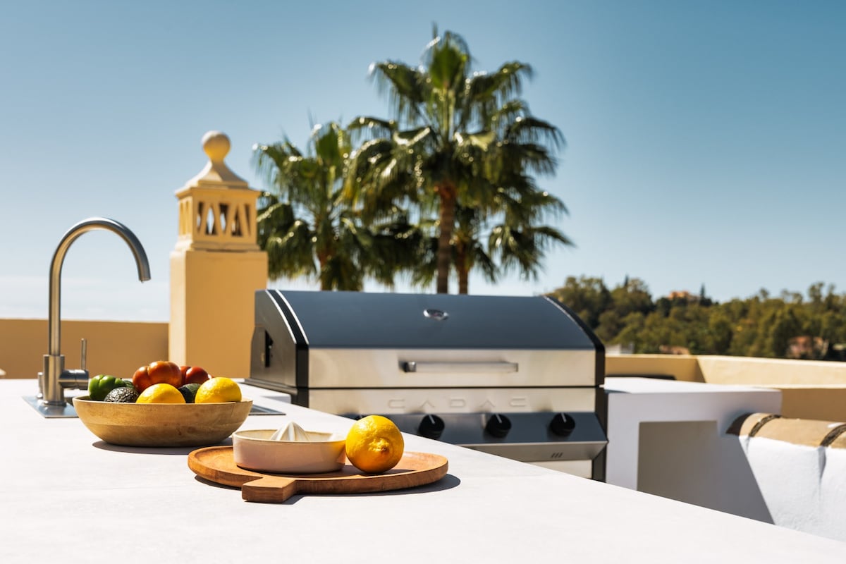 An outdoor kitchen area features a stainless steel barbecue grill next to a bar-style counter. Fresh fruits and herbs are displayed on a wooden board, while a modern sink is integrated into the counter. Lush palm trees are visible in the background under clear blue skies.