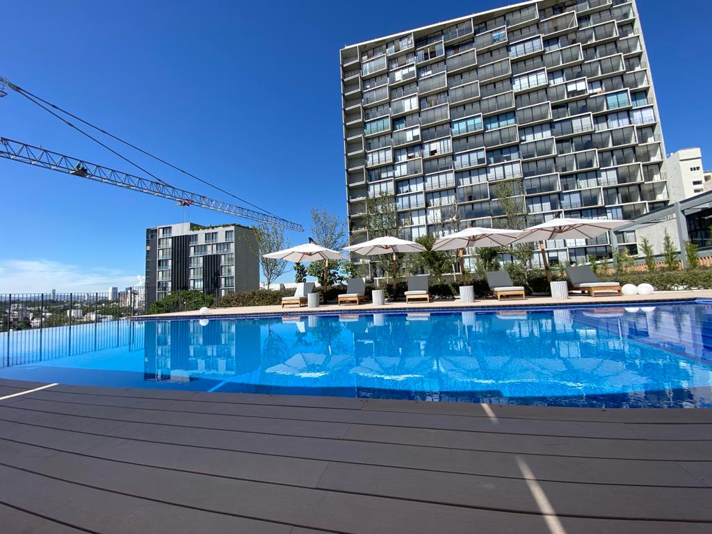 A modern swimming pool reflects the clear blue sky, framed by lounge chairs under white umbrellas. The surrounding landscaped area complements the sleek building in the background, showcasing a blend of contemporary architecture amidst greenery.