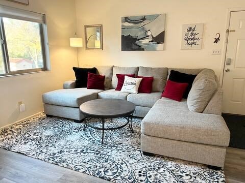 A comfortable sectional sofa in light gray is centered around a round coffee table. Decorative red and black pillows add contrast. A patterned area rug features shades of cream and gray. Natural light enters through a large window, with a piece of wall art visible nearby.