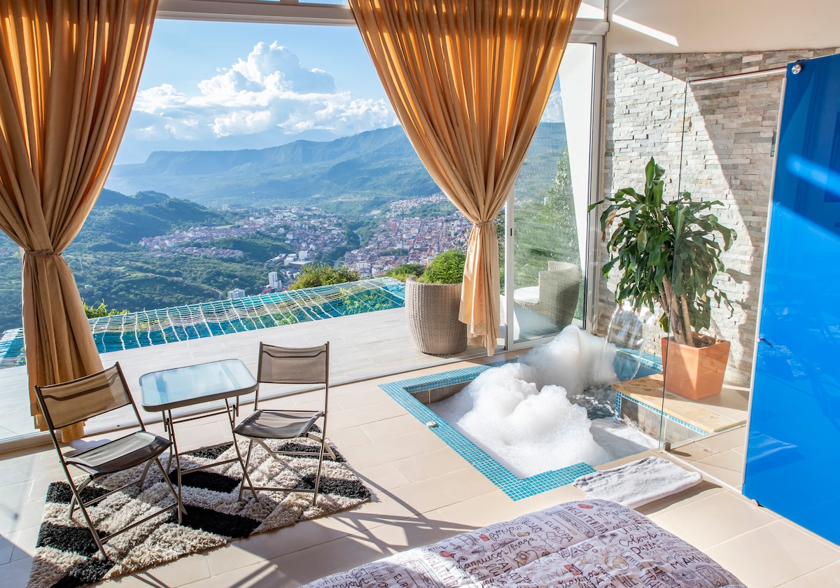 A spacious living area features large glass doors that open to breathtaking mountainous views. The room includes a cozy seating area with two chairs and a table, as well as a bubbling hot tub positioned beside a lush indoor plant.