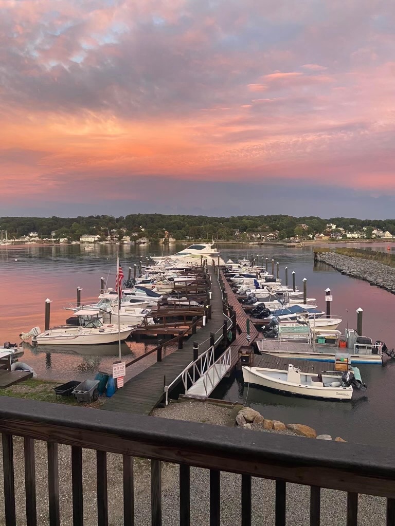 The image captures a serene marina at sunset, with colorful clouds reflected on calm waters. Several boats are docked along the wooden pier, illuminated by soft lights, creating a tranquil atmosphere. The waterfront is bordered by lush greenery and distant buildings, evoking a peaceful evening scene.
