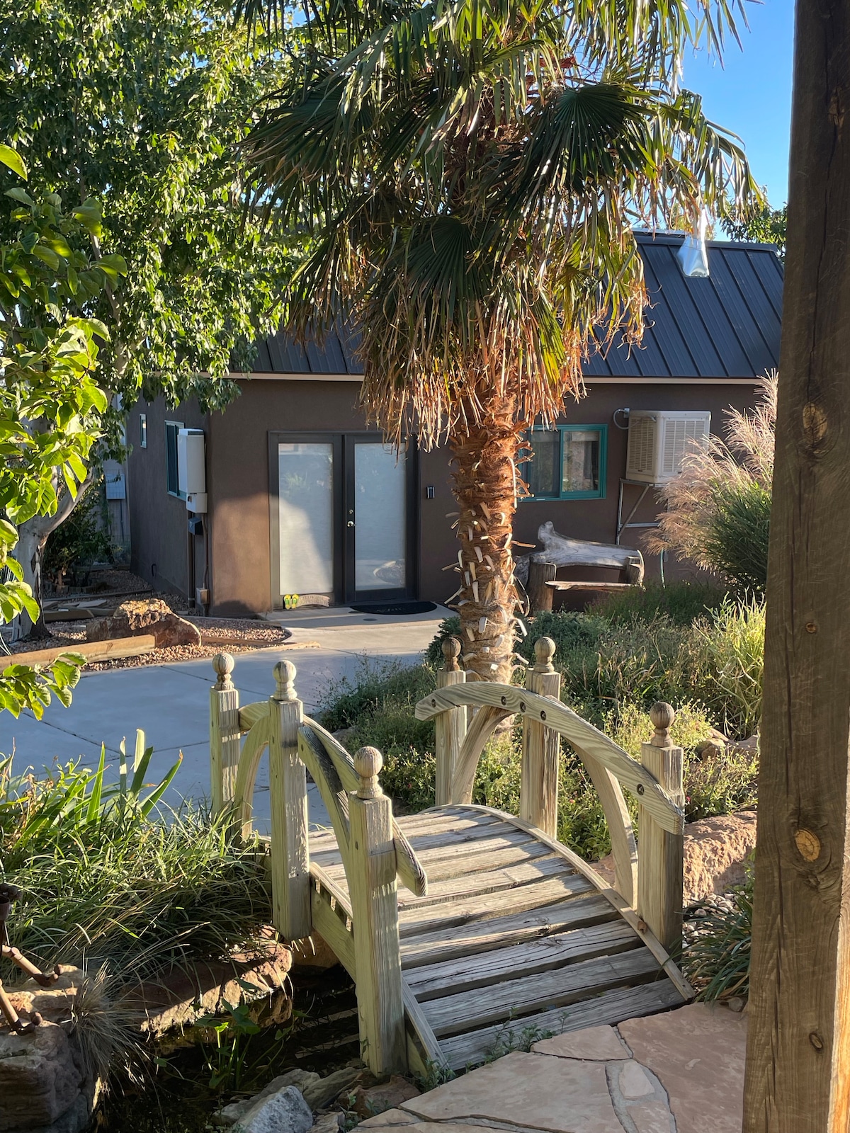 A wooden bridge arches over a small stream, leading into a landscaped area featuring various plants. In the background, a brown building with a metal roof is visible, along with a palm tree and a patio space.