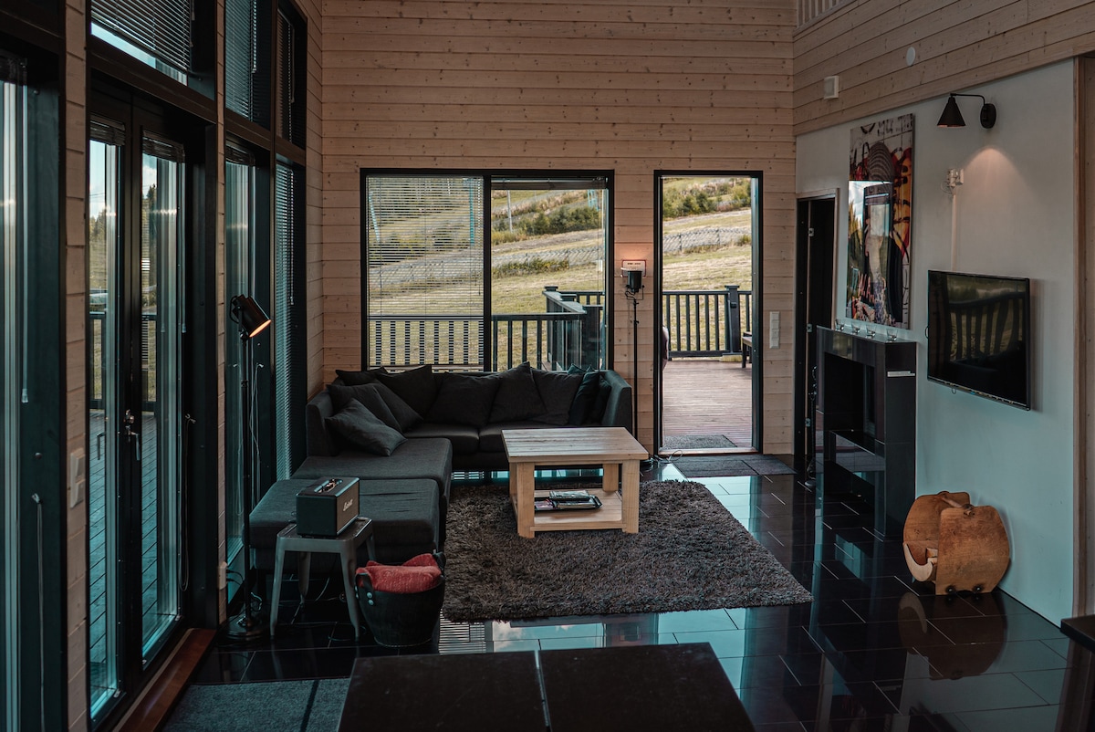 A spacious living area features large glass windows, providing views of the surrounding landscape. A cozy corner sofa faces a wooden coffee table, while a sleek TV is mounted on the wall. Dark flooring contrasts with light wood paneling, creating a modern yet warm ambiance.