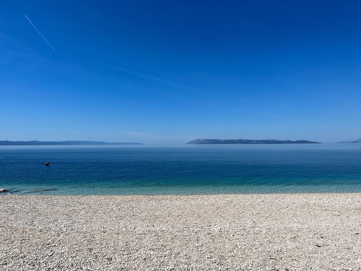 A tranquil beach scene features a smooth pebble shoreline meeting clear blue waters. The horizon stretches into the distance under a clear sky, with gentle waves lapping at the shore. The calming color palette includes shades of blue and turquoise, inviting relaxation.