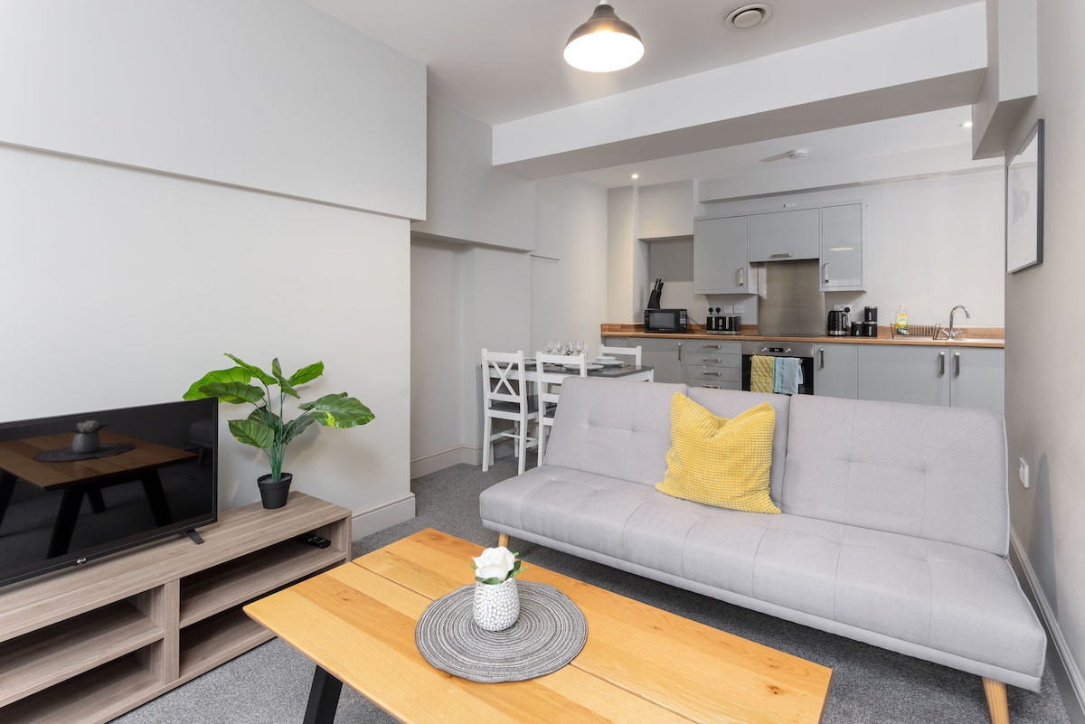 A modern living space features a light grey sofa bed adorned with a yellow throw pillow, positioned across from a television on a wooden stand. A wooden coffee table rests in front, while a dining area with white chairs is visible in the background, adjacent to a contemporary kitchen.