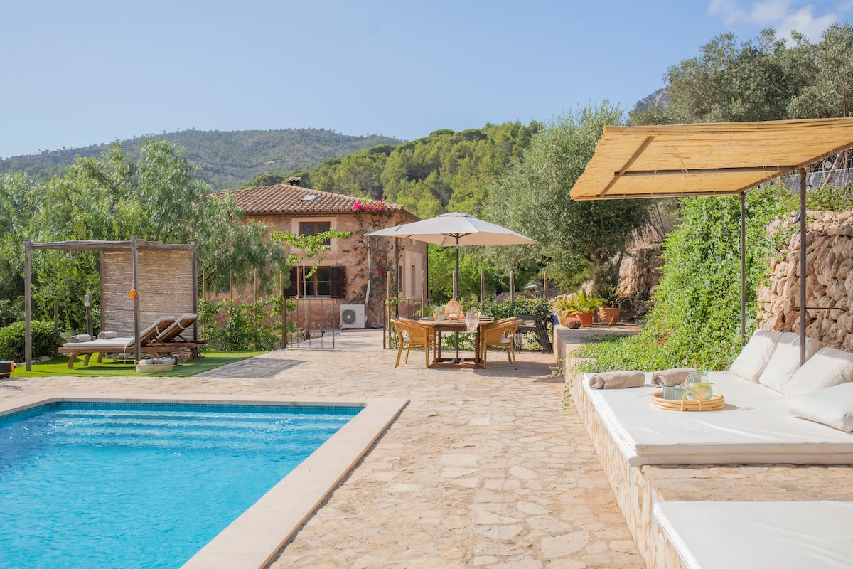 An outdoor area is showcased with a inviting swimming pool surrounded by natural stone tiles. Sunbeds are placed near the pool, while a shaded dining area features a table and chairs. Olive trees and greenery enhance the serene setting against a backdrop of mountains.