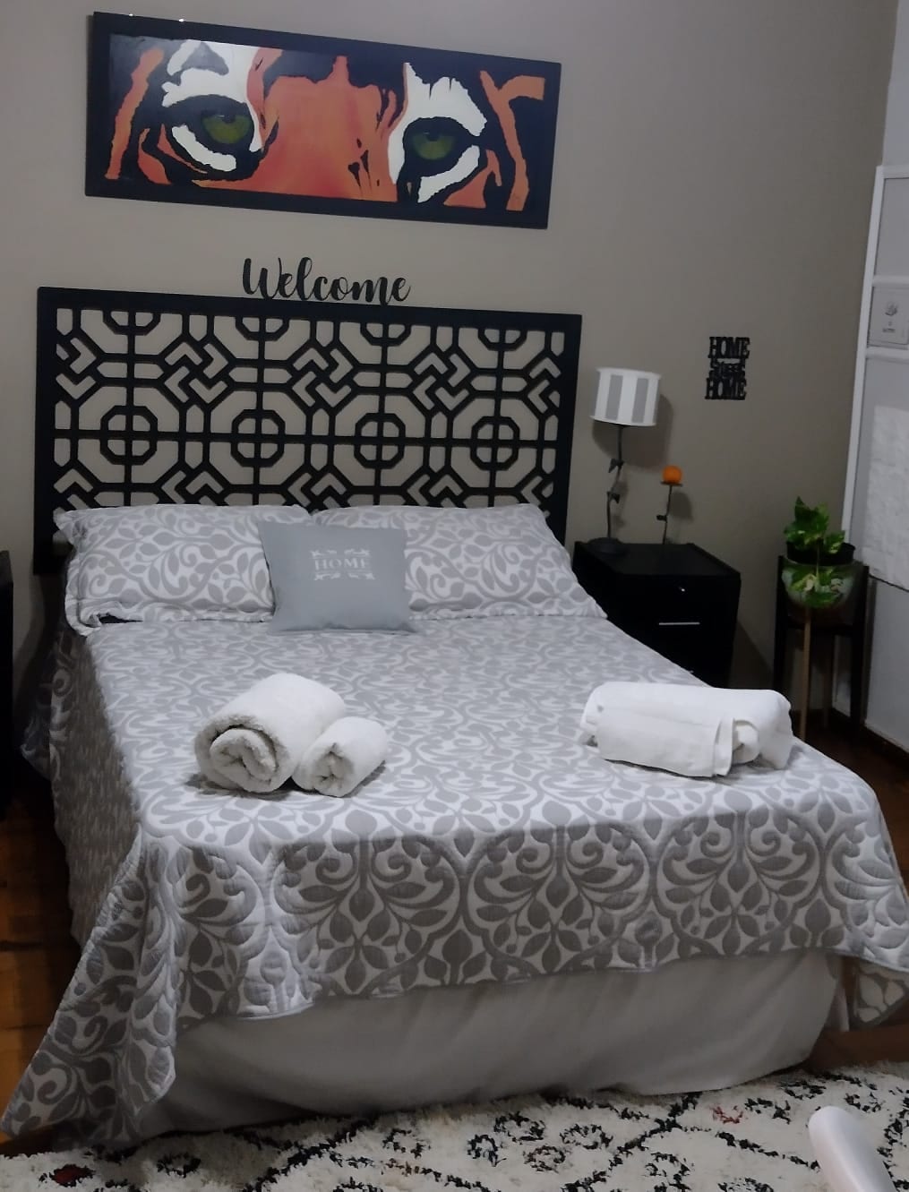 A cozy bedroom is highlighted by a patterned bedspread covering a spacious bed. Soft white towels are neatly arranged on the bed, and a decorative wall art piece featuring a tiger's face adds interest above the headboard. A bedside lamp and plant accompany a small nightstand.