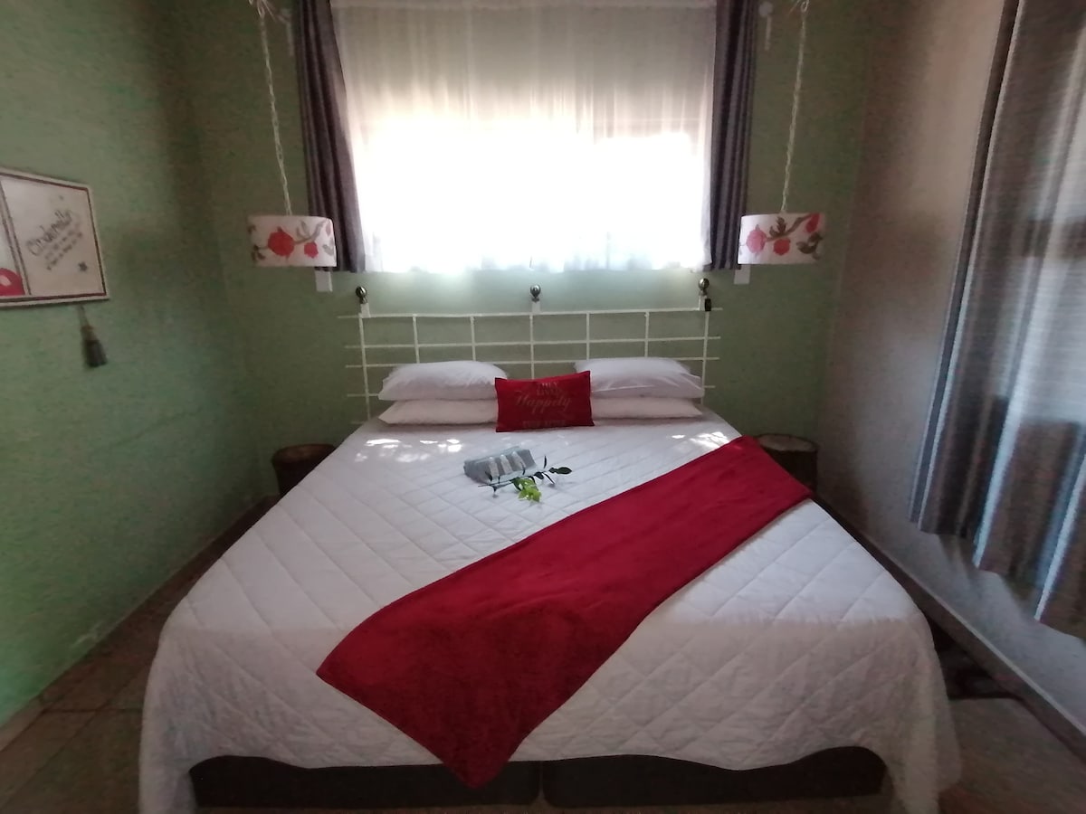 A cozy sleeping area features a spacious bed adorned with a vibrant red throw and decorative pillows. Soft natural light filters through sheer curtains, illuminating the green walls and pendant lights hanging on either side of the bed.