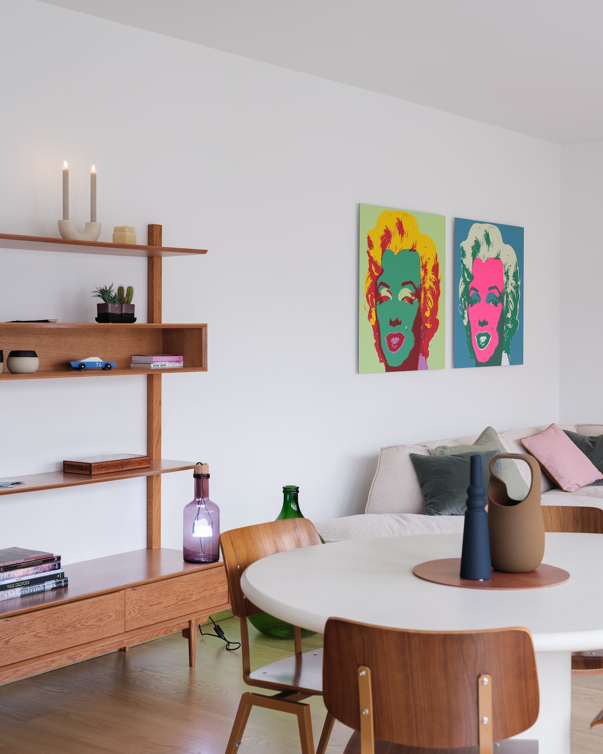 A modern living area features a dining table surrounded by wooden chairs. Two vibrant Marilyn Monroe prints adorn the walls. A wooden shelf displays decorative items and books, while a purple lamp adds subtle lighting to the space.