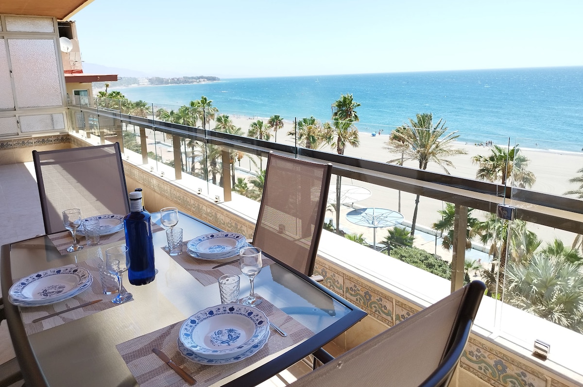 A spacious terrace features a dining table set for four, complete with plates and glassware. The view overlooks the beach and the Mediterranean Sea, framed by palm trees. Bright sunlight enhances the coastal scenery, creating an inviting space for outdoor meals.