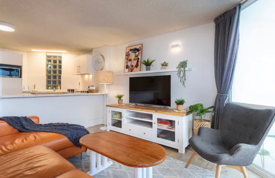 An open-plan living area is illuminated by natural light from sliding glass doors. A large, curved sofa features a cozy throw, and a flat-screen TV is positioned on a white console. Potted plants and artwork add vibrant touches to the modern decor.