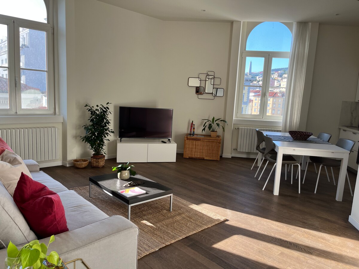 A spacious open-plan living area features a comfortable sofa and a smart TV. A dining table with chairs is presented alongside a decorative plant. Light floods the room through large windows, offering views of the surrounding cityscape.