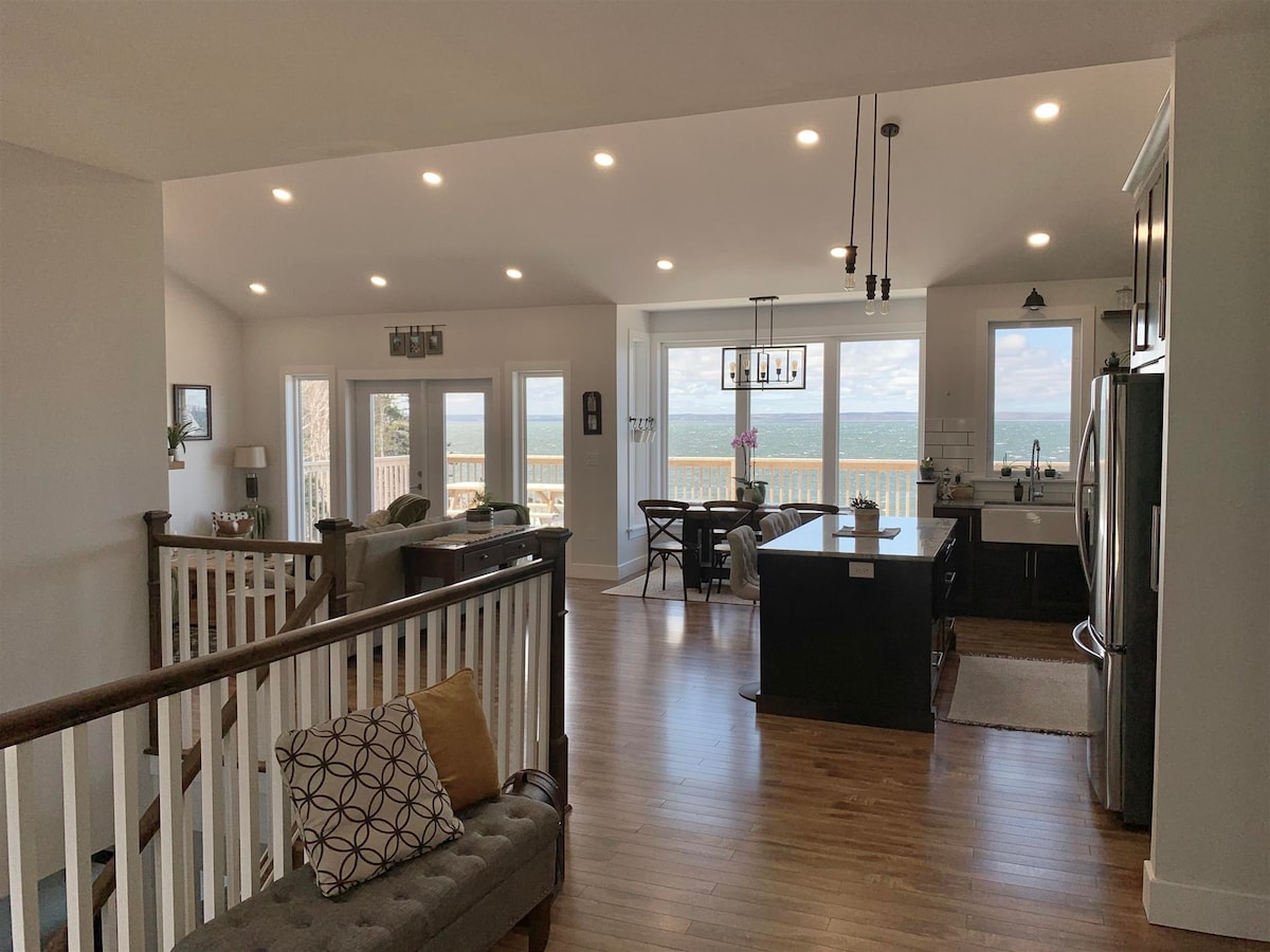 An open living space is showcased, featuring large windows that offer a view of the serene coastline. A modern kitchen with dark cabinetry is complemented by a dining area and inviting seating arrangements, illuminated by natural light streaming in from the expansive glass doors.