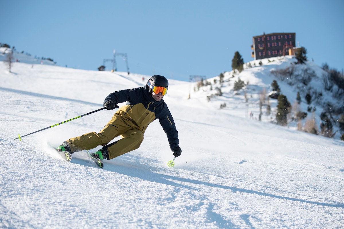 A skier navigates down a snowy slope, leaving trails in the fresh powder. In the background, a building is positioned atop the hill, surrounded by trees and ski lifts, with a clear blue sky above.