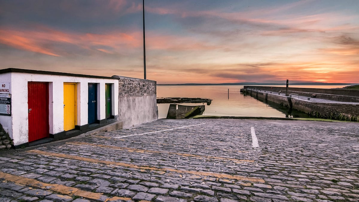 A serene waterfront scene is depicted, featuring a calm sea at sunset. A cobblestone area is visible, with brightly colored doors in red, yellow, and blue on a small building. The pier extends into the water, and soft hues fill the sky.