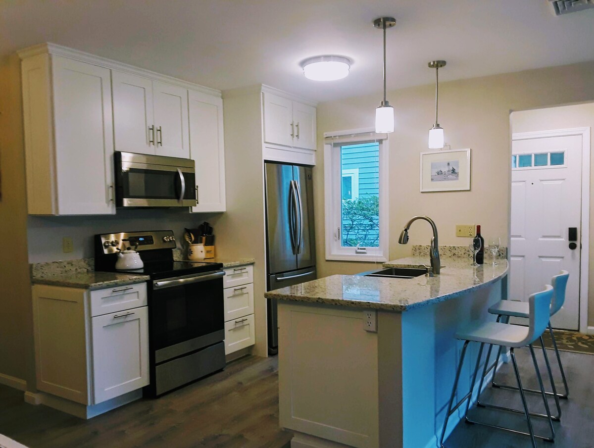 The modern kitchen features stainless steel appliances, including a microwave and refrigerator. Granite countertops provide ample workspace, complemented by white cabinetry. A kitchen island with seating is illuminated by pendant lights, and a window allows natural light to fill the space, with a door visible for access.