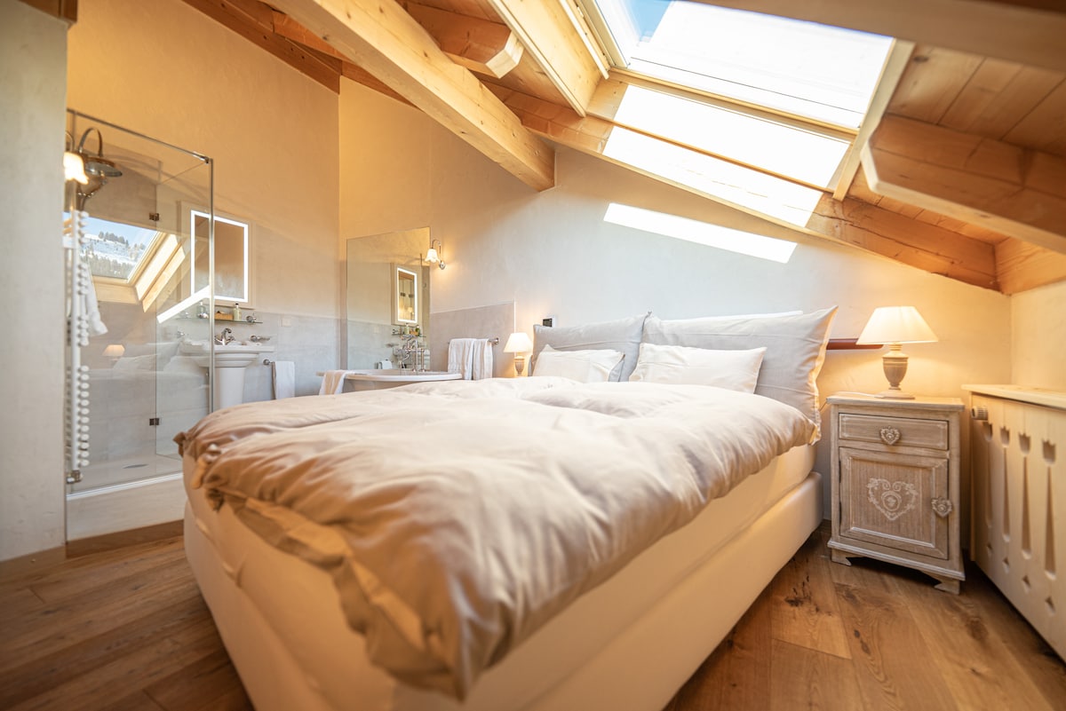 A cozy bedroom features a spacious bed with soft bedding, complemented by wooden beams along the ceiling. A large skylight offers natural light, while an attached open bathroom area is visible through glass panels. A bedside table with a lamp adds functionality to the space.