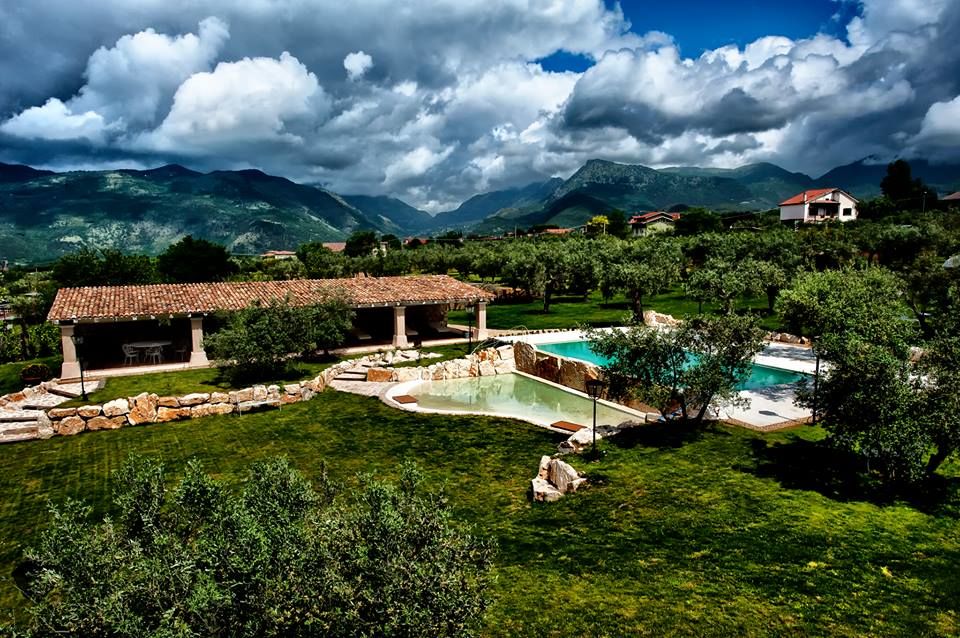 A scenic view depicting a lush landscape featuring olive trees and two swimming pools. The spacious grounds are complemented by a shaded terrace area with a tiled roof. Mountains rise in the background under a dynamic sky, presenting an expansive natural setting.