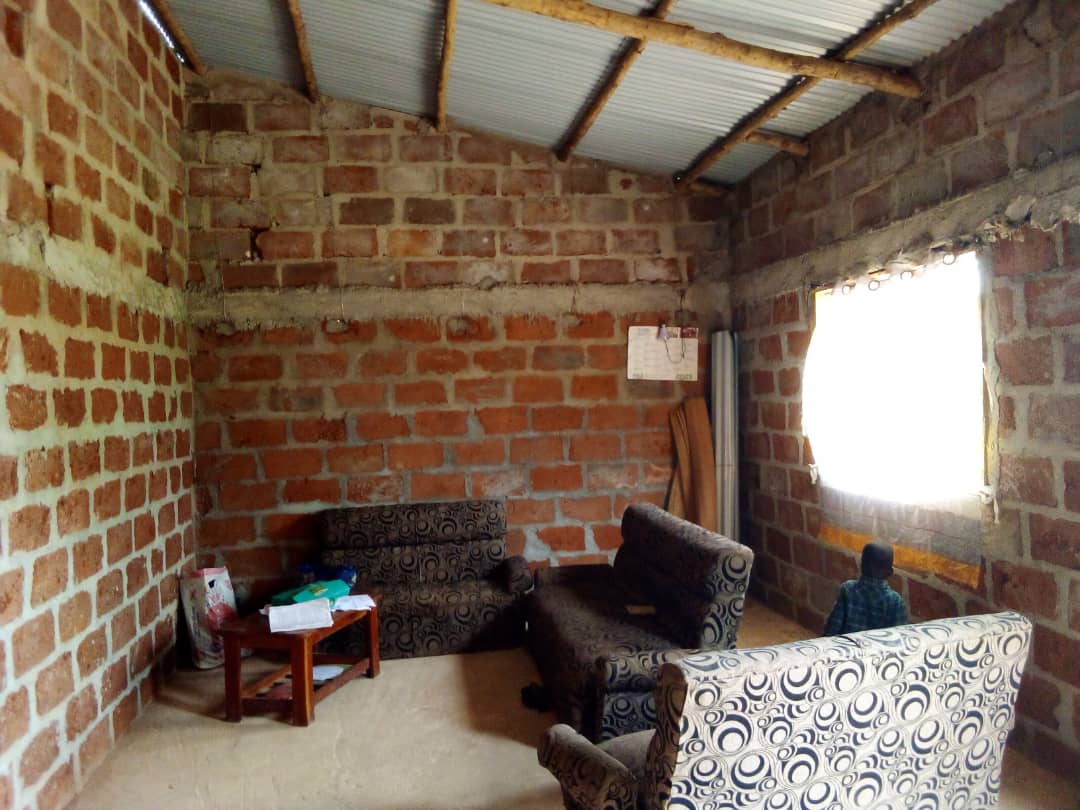 The living area features two patterned chairs and a small wooden table. Exposed brick walls create a rustic ambiance. Natural light enters through a window with sheer curtains. A few items are organized on the table, adding a subtle touch to the space.