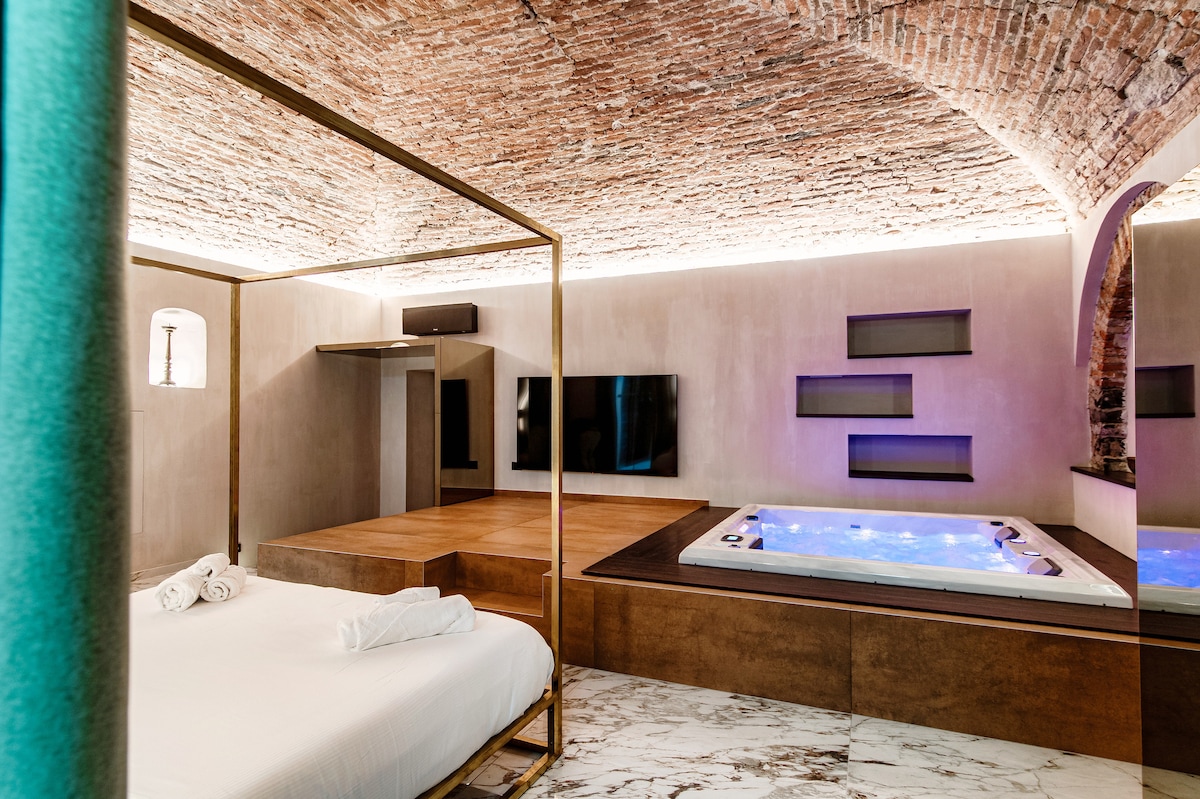 An open space is showcased with a luxurious golden canopy bed in the foreground. A sophisticated jacuzzi is positioned on an elevated platform to the side. Exposed brick arches and soft lighting contribute to the modern design, while light marble flooring enhances the ambiance.