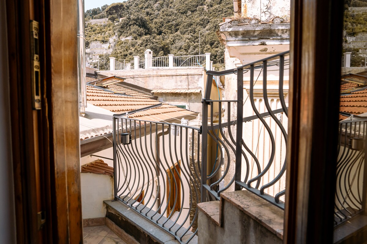 A view from a window shows a metal railing balcony overlooking neighboring rooftops and greenery. The architecture features rustic tiles and varying heights of surrounding buildings. Soft morning light illuminates the scene, highlighting the textures and colors of the structures.