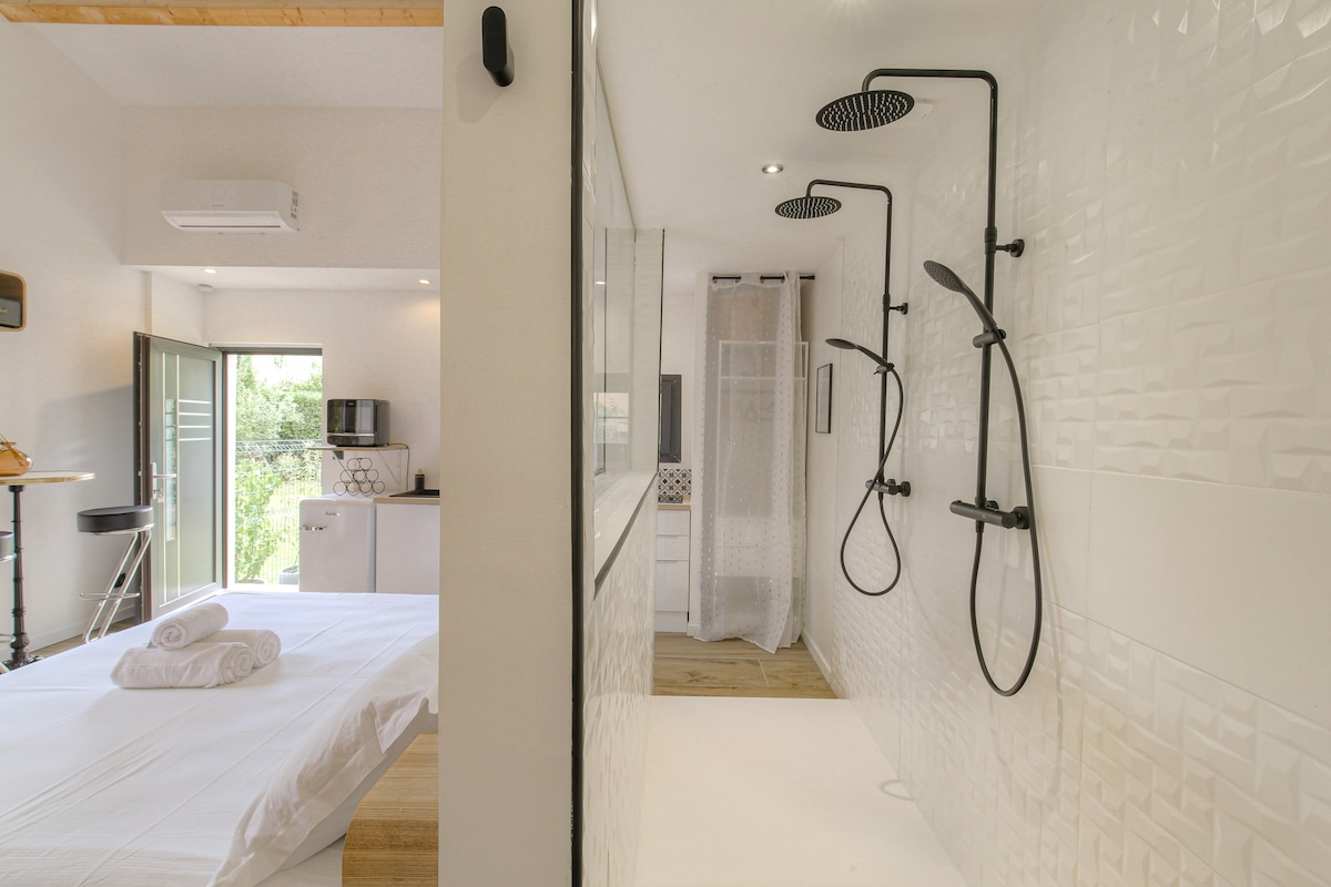 A modern bathroom space is showcased, featuring dual overhead rainfall showerheads. The sleek design is complemented by patterned wall tiles and a bright, airy ambiance. A doorway leads to a separate area with natural light, revealing a bedroom and kitchenette.